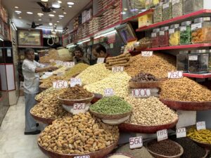 A shopkeeper stands behind a large display of nuts, dried fruits, and spices at Khari Baoli, Asia’s largest spice market, with many colorful bowls labeled with prices.
