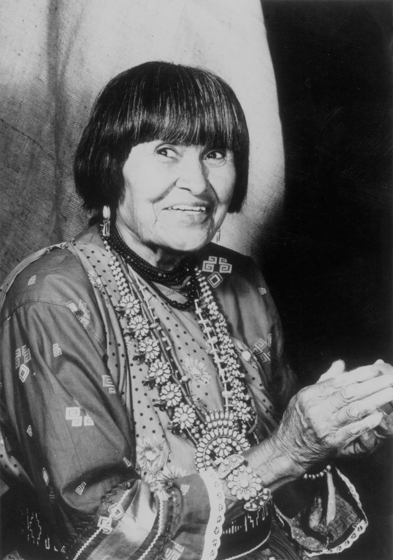 Black-and-white portrait of Pueblo potter Maria Martinez shows an older, Native American woman with short dark hair trimmed neatly around her face. She wears traditional, formal-looking clothing and beadwork jewelry, including an ornate necklace and dangling earrings. Her hands are clasped in front of her and she is looking slightly to the left of the camera, and smiling.
