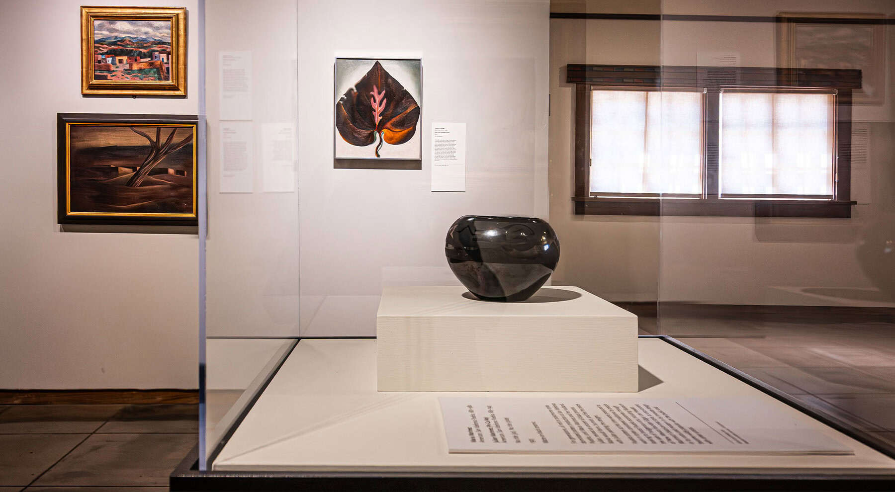 A blackware Maria Martinez ceramic pot shown on a white pedestal in a glass display case at a museum. Several framed paintings, including a Georgia O'Keeffe painting, hang on the walls in the background.