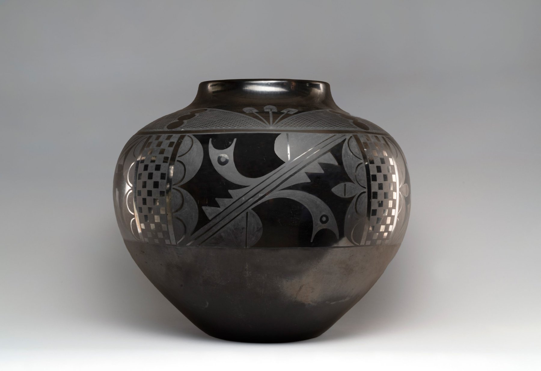 A round, black ceramic Maria Martinez black-on-black olla pot with a narrow opening and intricate, matte and glossy geometric and abstract patterns etched around the upper body. The background is plain and light gray.