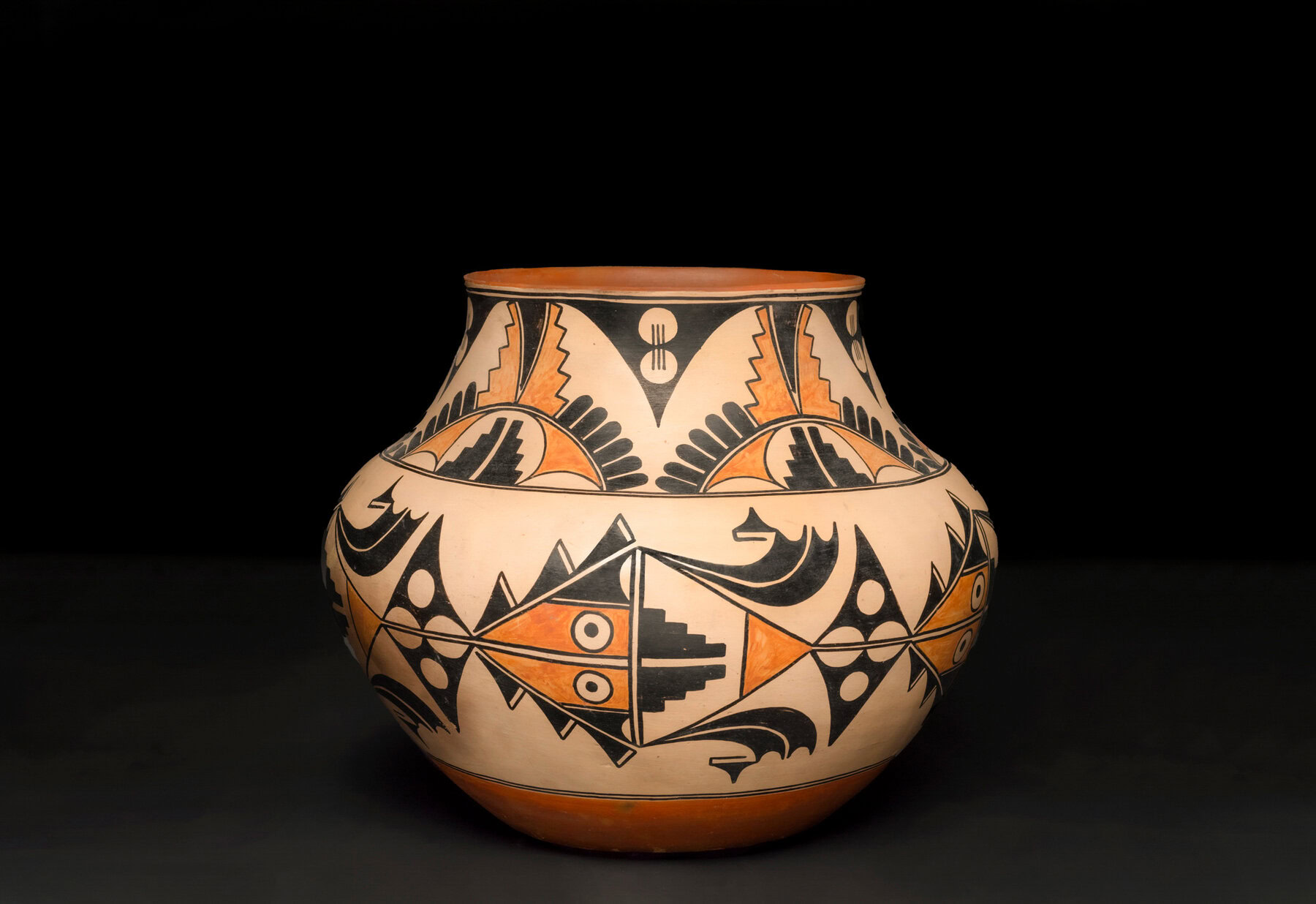 This ceramic polychrome pot made by Maria and Julian Martinez has an inverted bulb shape, and is decorated with geometric and abstract patterns in black and orange on a cream background. The pot is photographed against a black backdrop.
