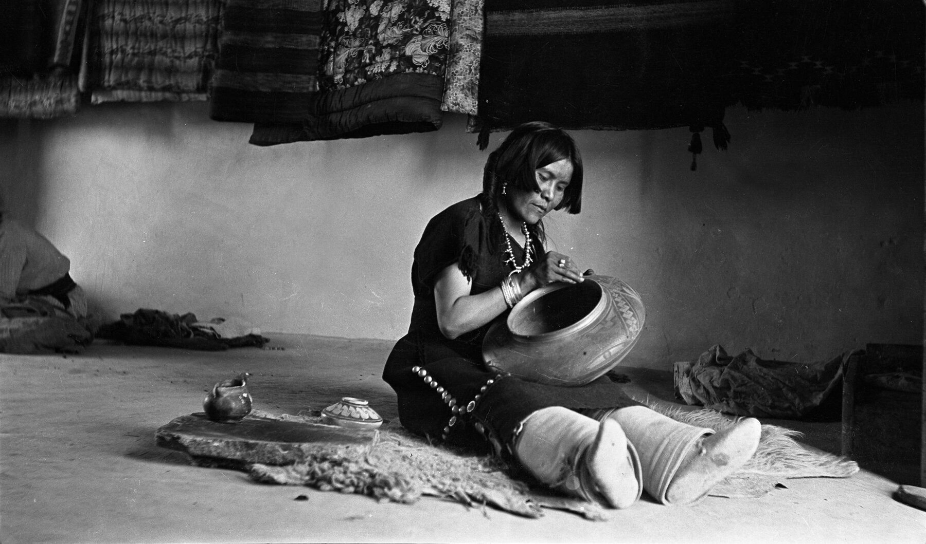A woman, believed to be the famed Hopi potter Nampeyo, sits on a rough floor holding a large clay vessel in her lap. She wears traditional clothing and jewelry, and is surrounded by woven blankets and other pieces of pottery in a softly lit indoor space.