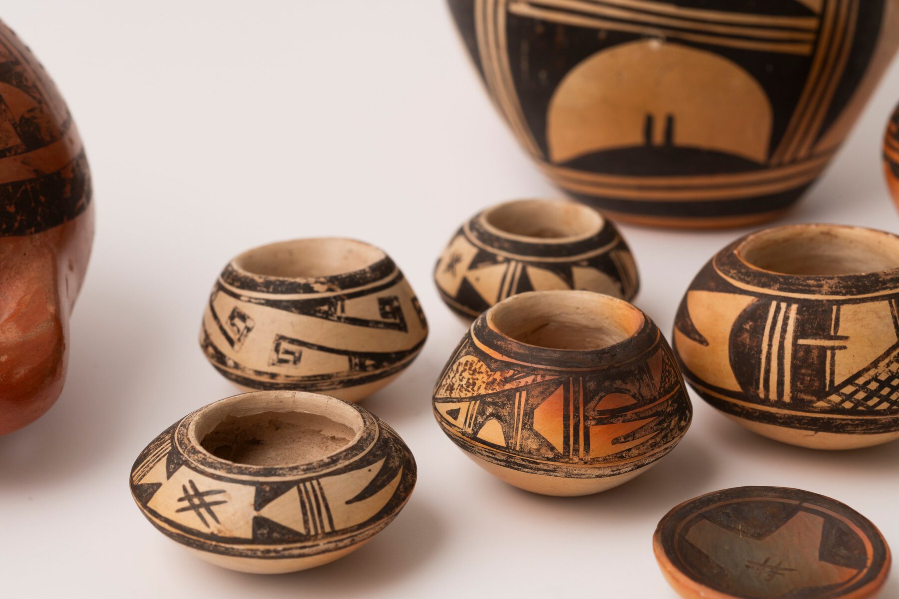 Several small, round miniature ceramic pots made by Nampeyo and painted in her signature Sikyatki style, featuring intricate geometric and abstract earth-tone designs. The pots are arranged on a white surface, with parts of larger pottery pieces partially visible in the background.