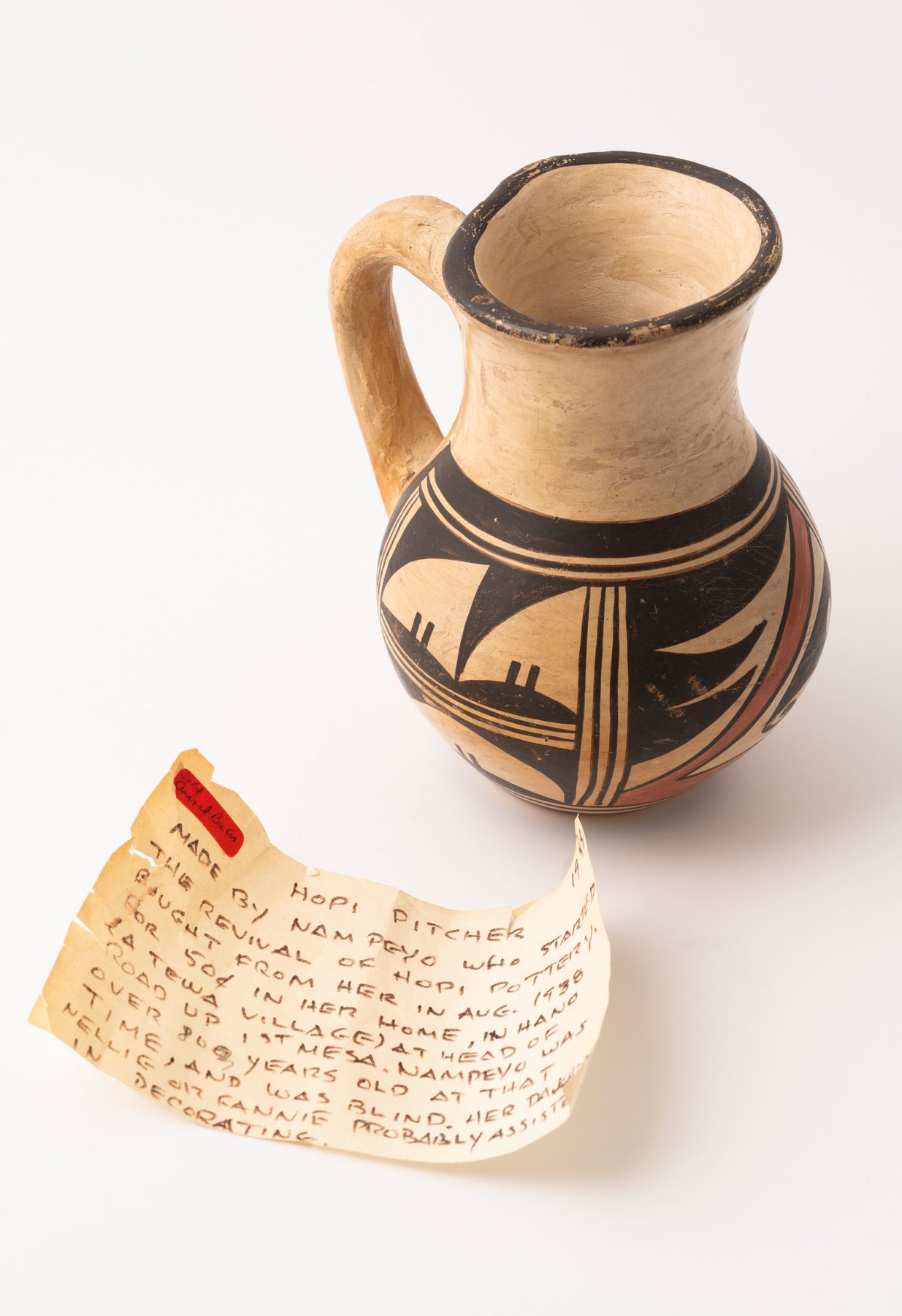 An antique, handmade ceramic pitcher with geometric designs and showing signs of age, sits beside a handwritten note that describes its origin as a Hopi pitcher made by the potter Nampeyo and possibly decorated by one of her daughters.