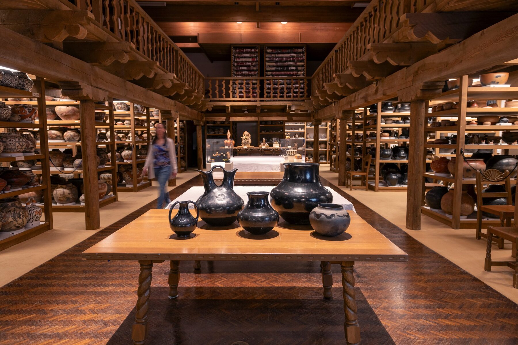 A large room with warm, low lighting shows several rows of wooden shelves holding a large collection of Southwestern style pottery, vases, and artifacts. In the foreground, a wooden table holds a collection of pots made by the Pueblo potter Serafina Tafoya. A blurred female figure can be seen in the background on the left, apparently walking toward the camera.