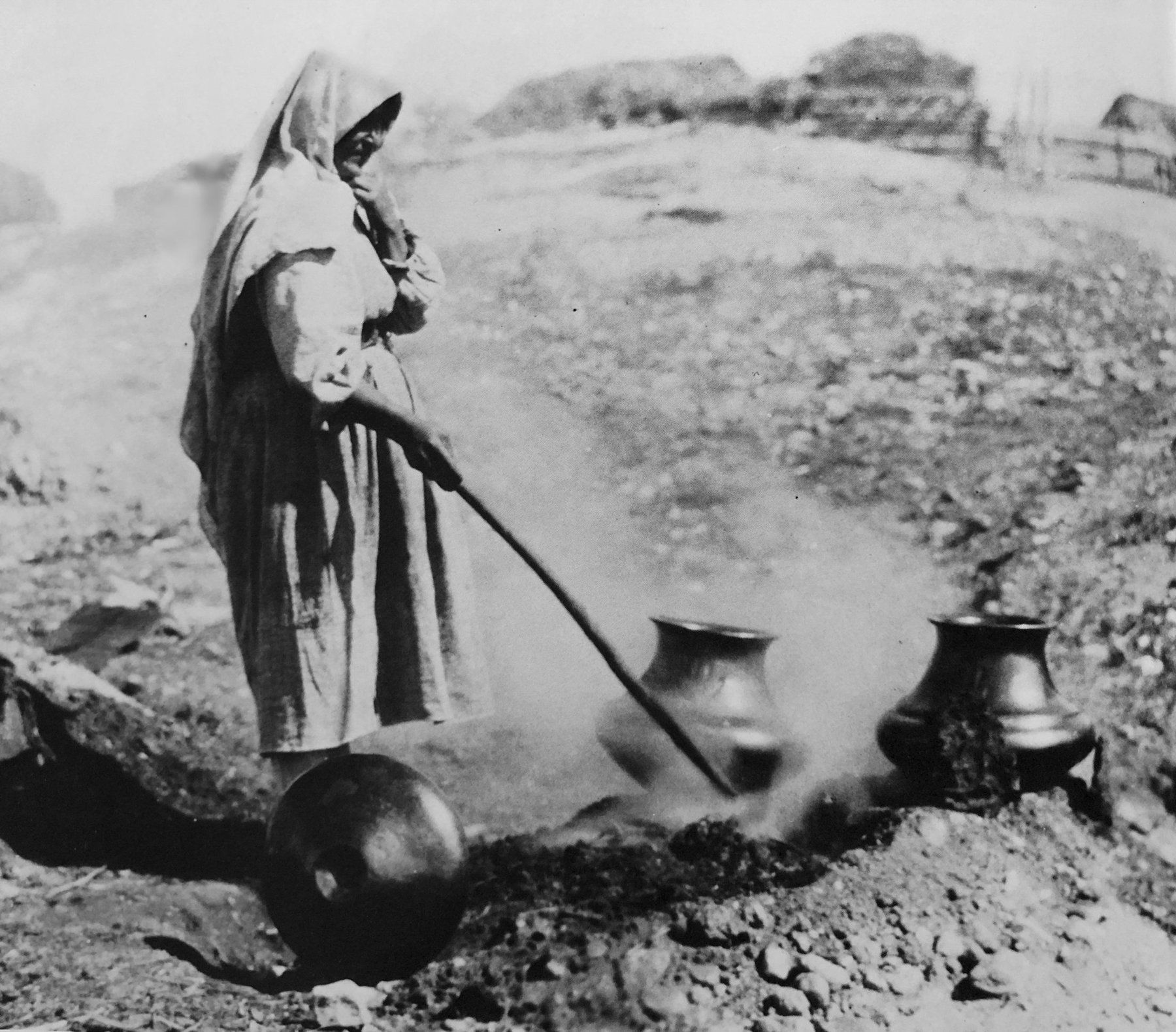 A black-and-white image of a female figure standing outdoors, tending to three large ceramic pots over a smouldering fire on the ground. Smoke is rising around her and the background appears as a blurred, rocky landscape with what may be part of a building roofline and a fence on the horizon.