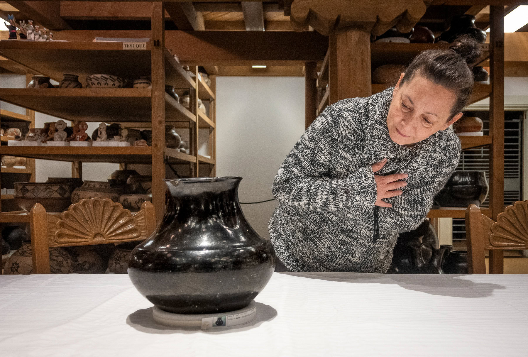 The author of this story, RoseMary Diaz, who is Serafina Tafoya's great-great-granddaughter, leans in to examine a large, black ceramic pot made by her ancestor. The pot is displayed on a light-colored table. The woman is wearing a patterned black-and-white sweater and her dark hair is pulled back; one hand rests expressively below her throat. Behind her are several wooden shelves full of pottery.