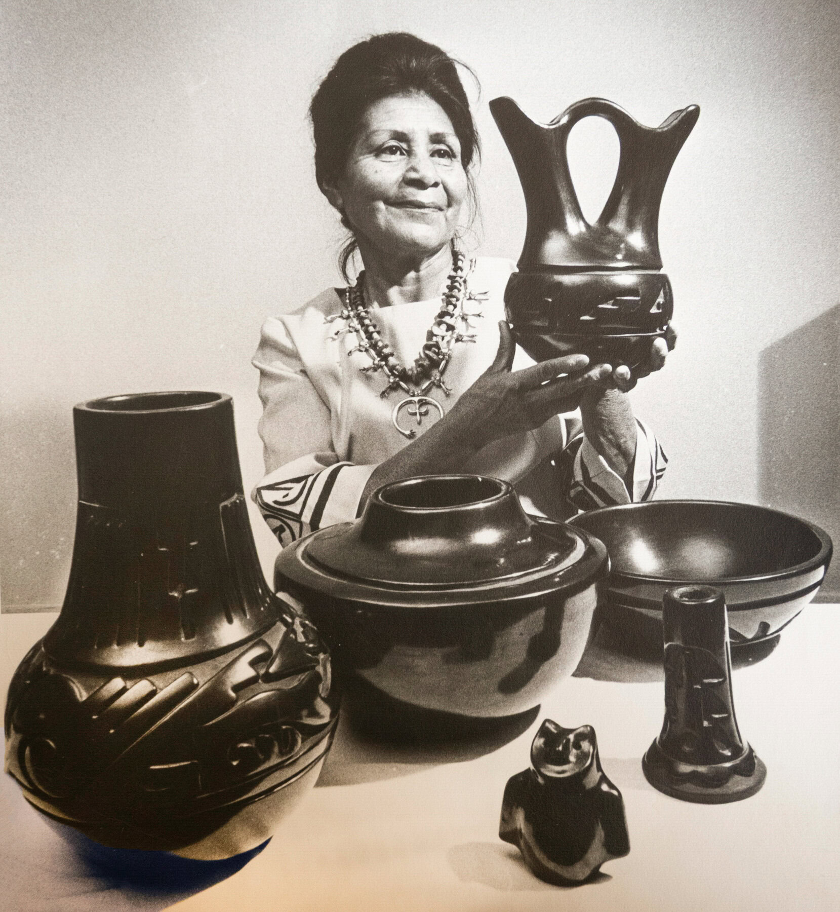 A black-and white portrait shows a seated woman with dark, upswept hair wearing an ornate beaded necklace and a traditional Pueblo-style dress. She holds up a sculpted and carved vase she has made. In the foreground are several other black pottery pieces, including vases, bowls, and a small animal figurine, arranged on a table.