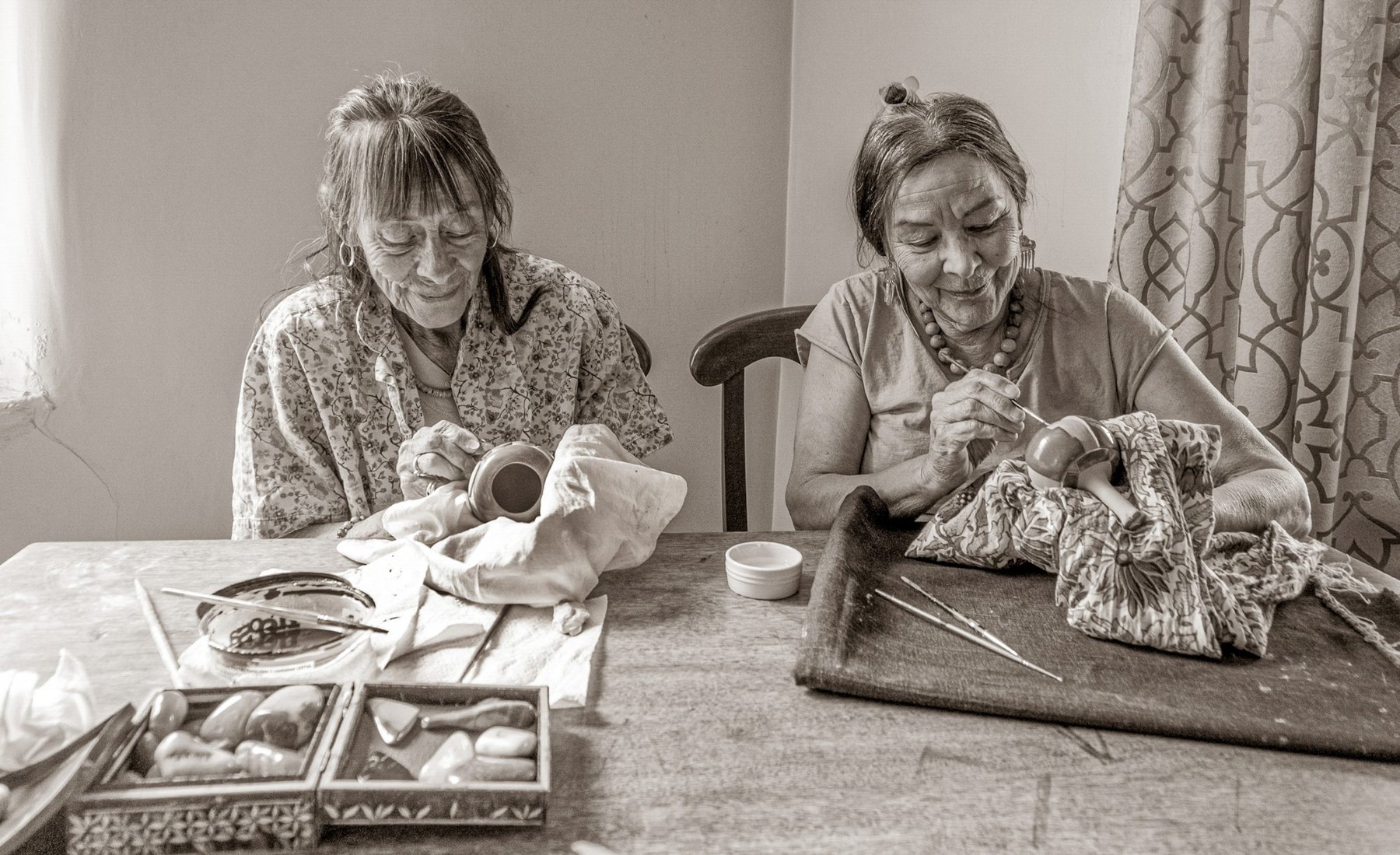 Two elderly women sit at a table, carefully painting pottery pieces by hand. Various tools and brushes are scattered on the table, and the women appear focused on their detailed work.
