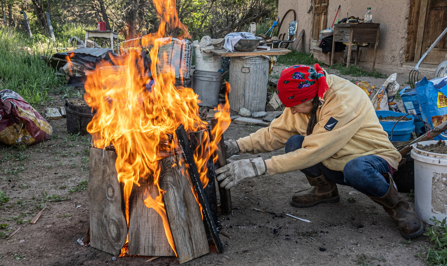 An elderly female figure wearing heavy work gloves, boots, and a bright red scarf on her head tends to a tall, blazing wood fire outdoors. She is surrounded by buckets, tools, and various objects on the ground. A rustic adobe building can be partially seen in the background.