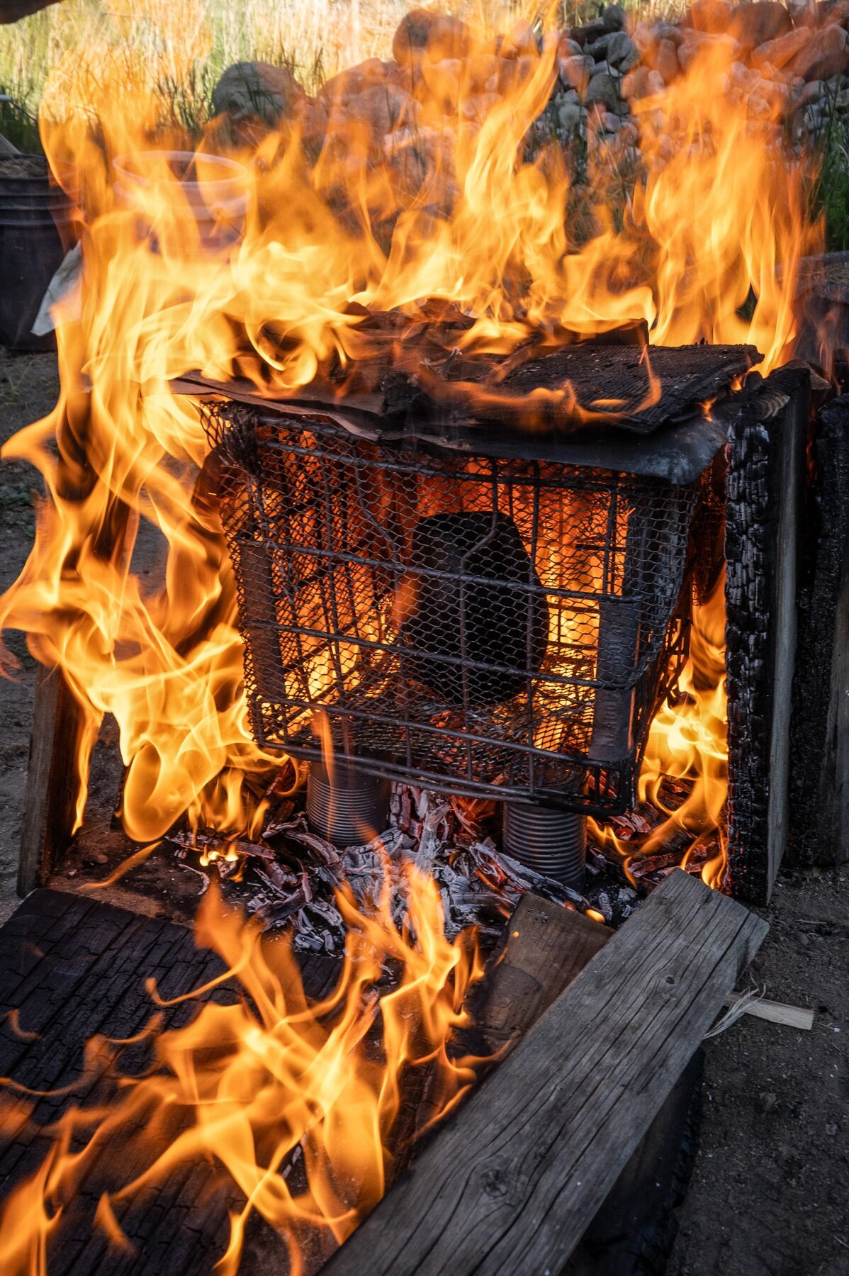 A metal wire crate containing a dark, rounded ceramic pot is propped up by tin cans and engulfed by bright orange flames, burning wooden planks, and embers. Rocks and grass are visible in the top of the frame, as a blurred background.