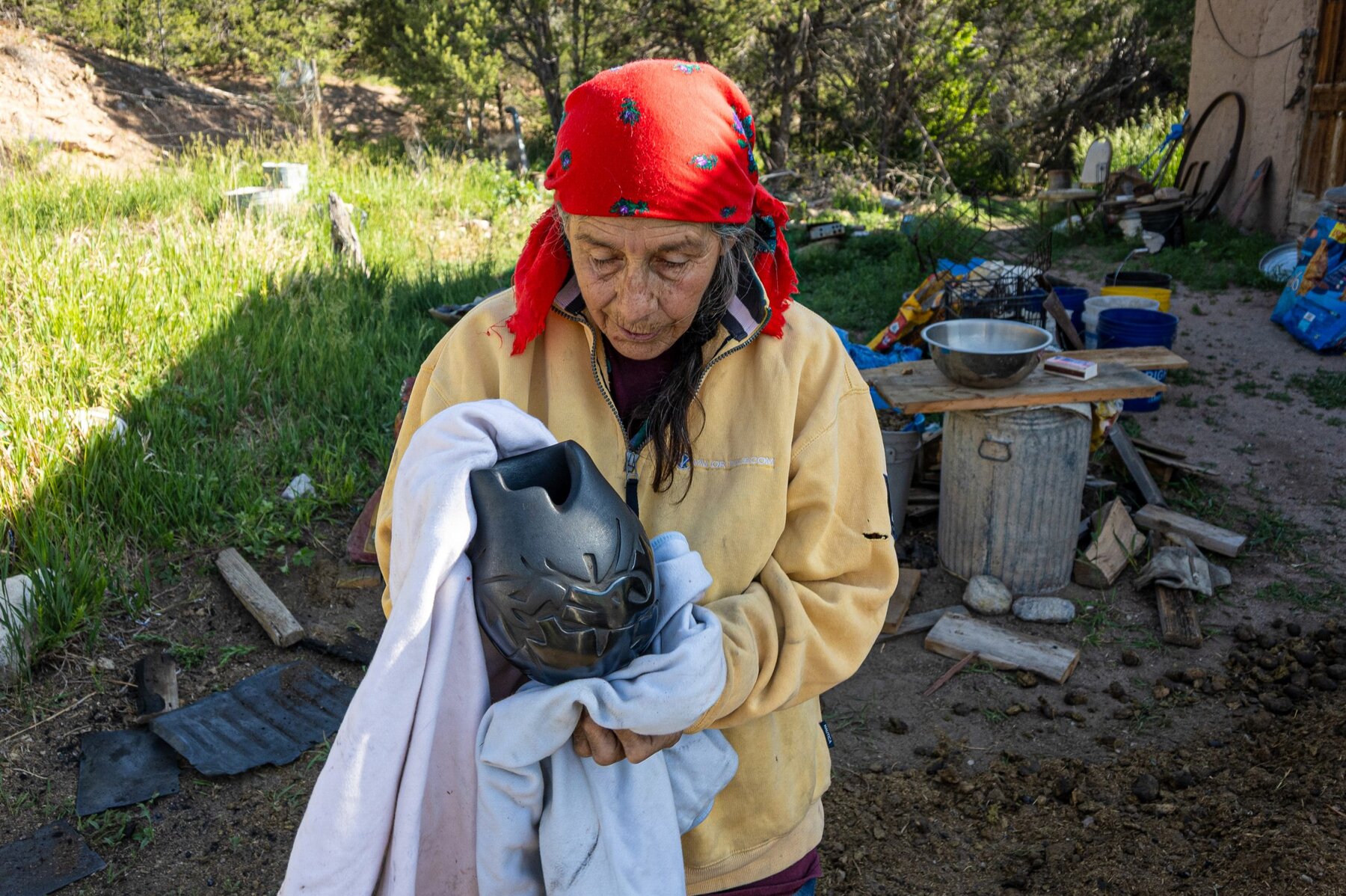 An elderly woman in a red headscarf and yellow fleece jacket holds a lustrous black pot carved with a design and partly wrapped in a cloth. She is standing outdoors surrounded by rustic tools, overgrown grass, and a makeshift wooden table. A few planks of unused firewood are scattered about on the ground.
