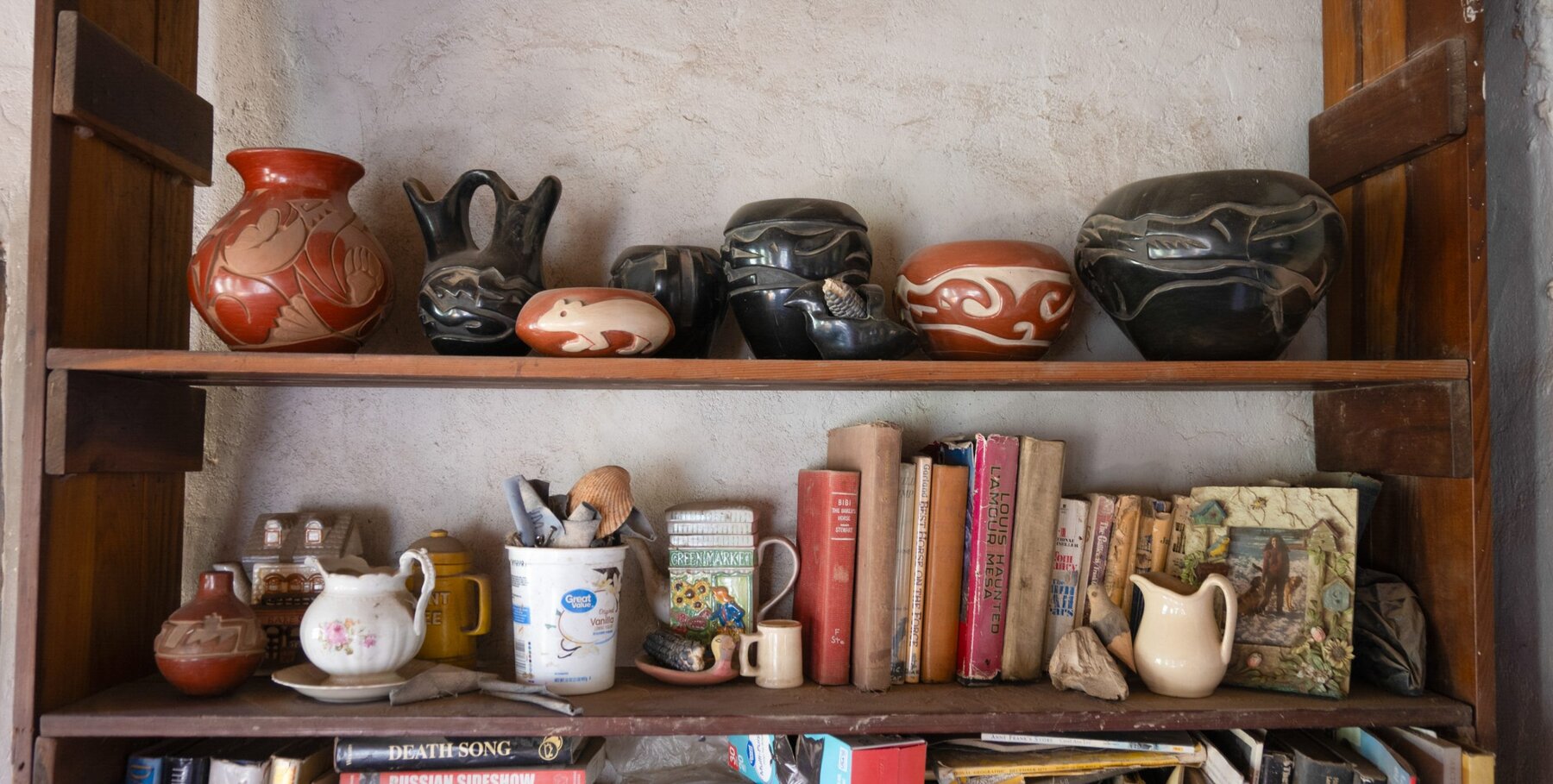 A rustic wooden shelf displays a collection of Pueblo pottery on the top, and assorted books, teapots, tins, and small figurines on the lower shelf against a light-colored adobe wall.