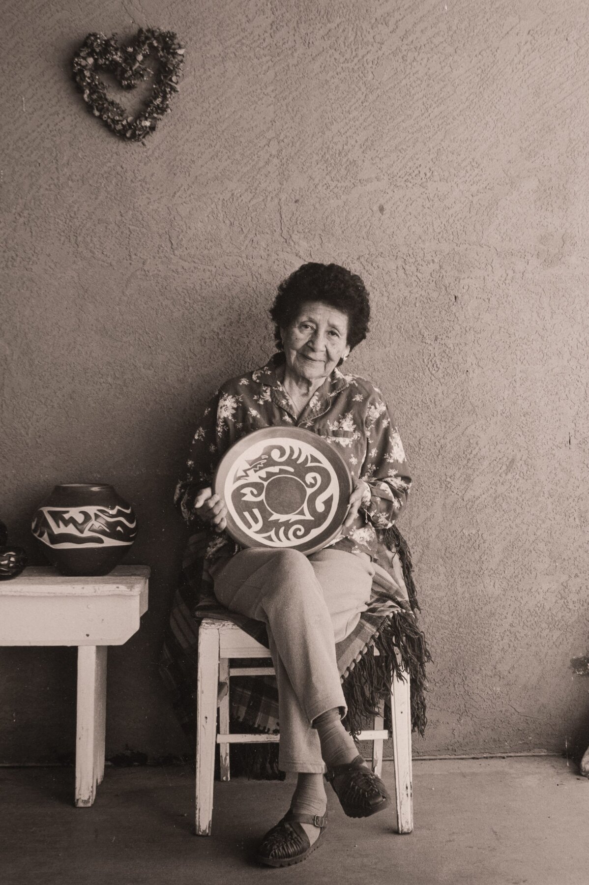 A sepia-toned portrait of elderly woman seated on a chair against a textured wall, holding a ceramic plate with a boldly carved design. A pottery jar rests on a small table nearby, and a heart-shaped wreath hangs on the wall above her.