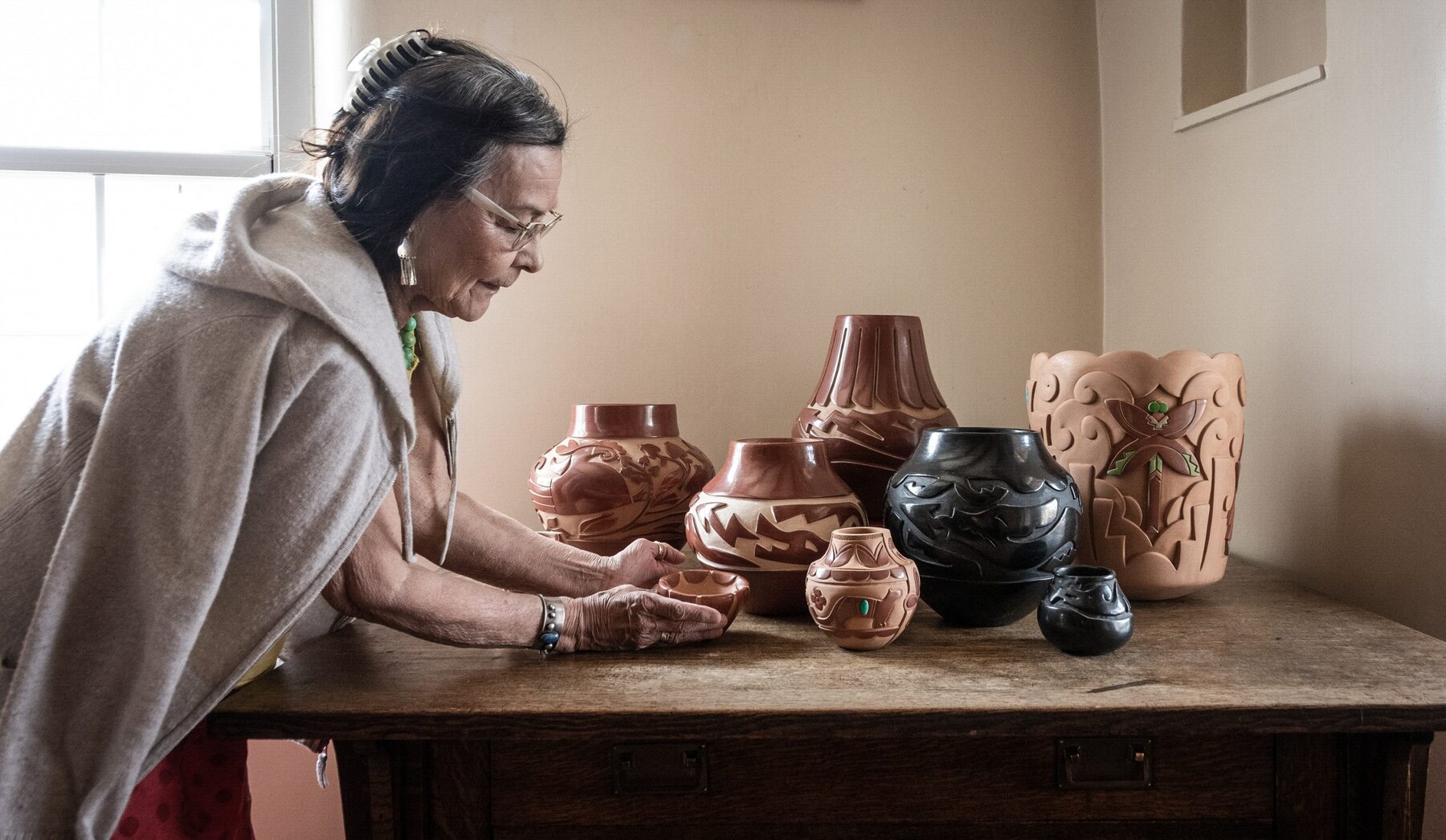 A woman wearing glasses and a shawl arranges a collection of intricately decorated pottery on a wooden table in a softly lit room. The pottery varies in size, shape, and color.