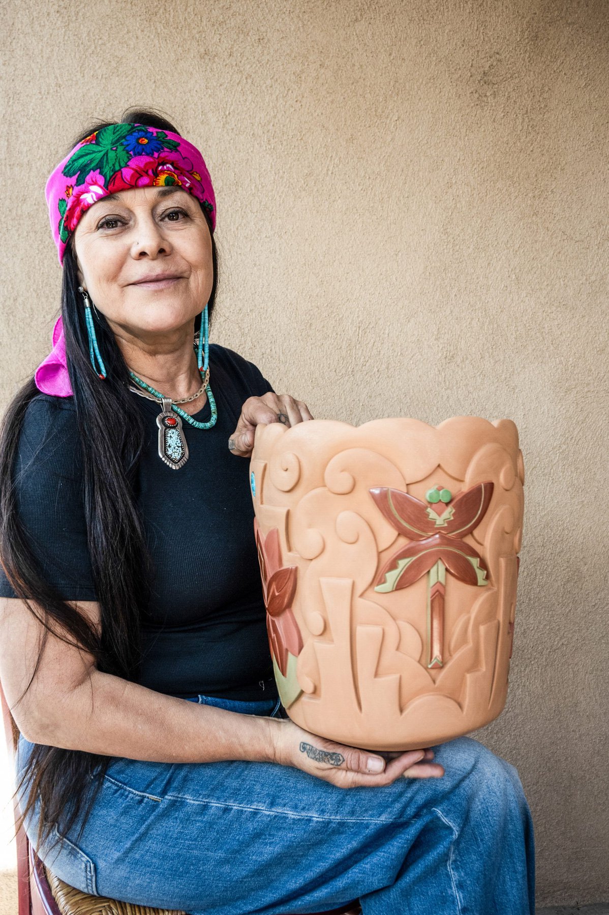 A seated woman with long dark hair, wearing a colorful embroidered headband, black shirt, blue jeans, and beaded jewelry, holds a large, decorative clay pot she has made and carved with a stylized dragonfly design. In the background is a light-colored wall with some texture, like adobe.