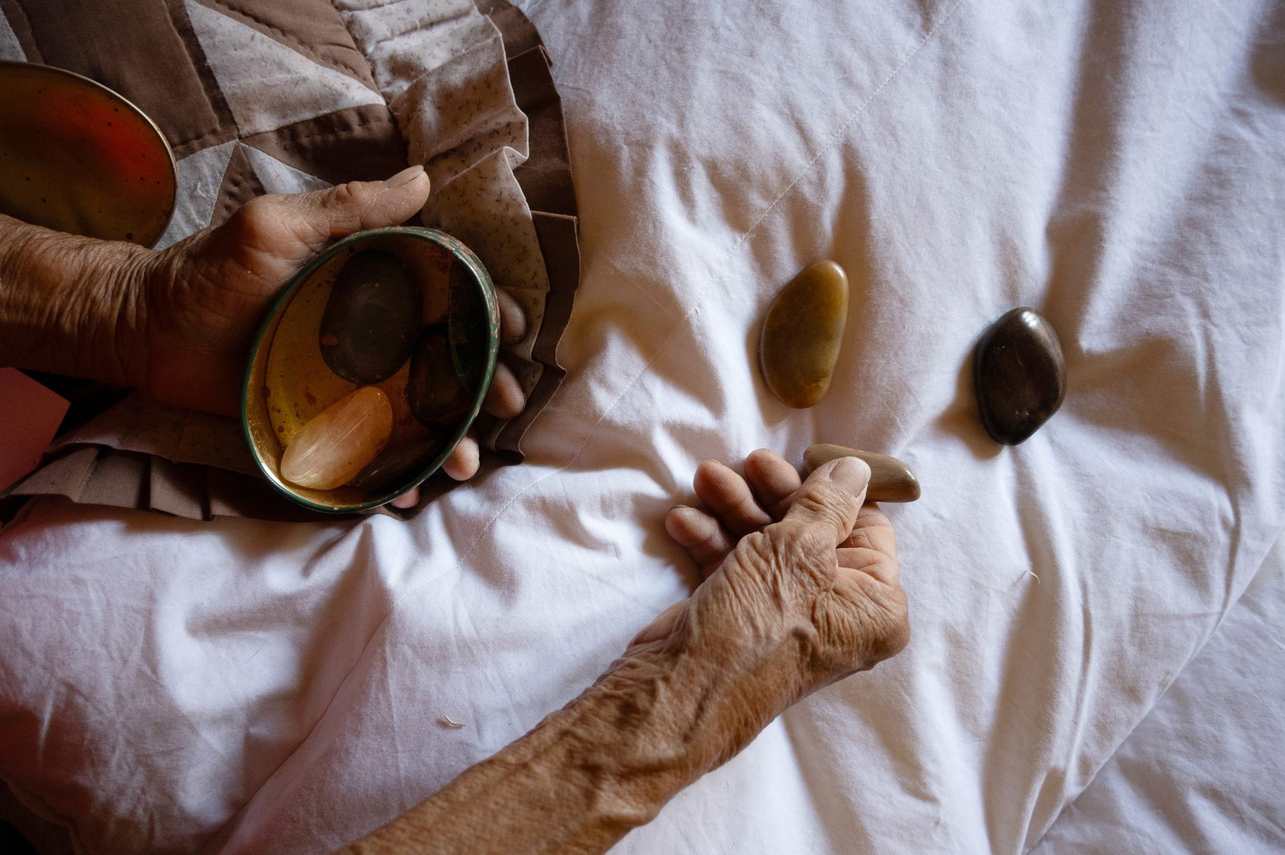 A pair of elderly hands arranges a few smooth, polished stones on a white blanket, holding a small bowl with more stones in one hand.