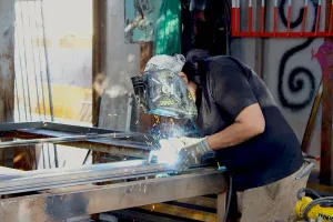 A person wearing a welding helmet and gloves is welding metal pieces together on a workbench, with bright sparks visible. Workshop tools and equipment are visible in the background.