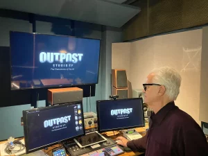 A man with white hair and glasses works at a sound mixing desk in a studio. Multiple monitors display the OUTPOST STUDIOS SF logo. Audio equipment and speakers fill the room.