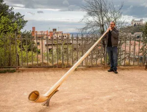 A man plays a large alphorn in an outdoor area, with buildings and a hilltop church visible in the background under a cloudy sky.